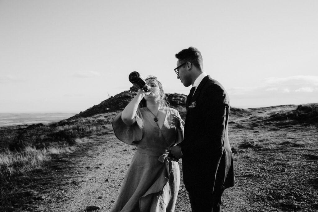 bride and groom on hilltop with bride drinking from bottle of champagne