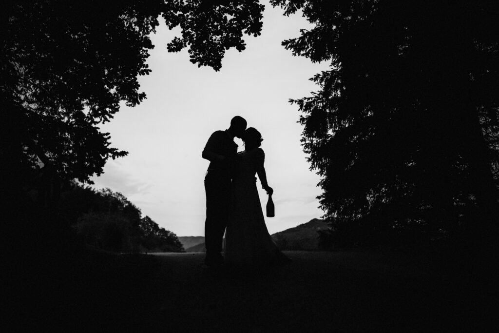 bride and groom silhouetted in trees bride holds bottle of champagne